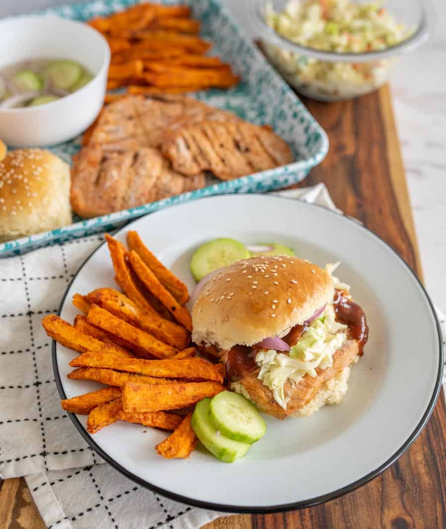 A white plate with a grilled Pork chop sandwich, cucumbers, and sweet potato fries with the serving tray of all those foods in the background.
