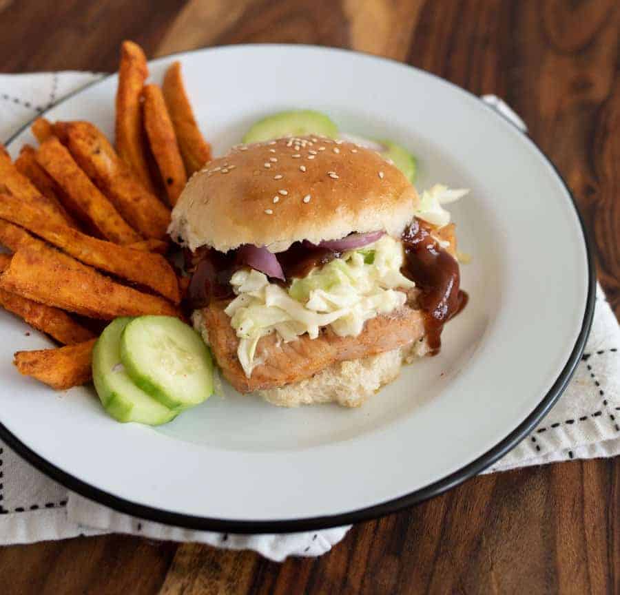 A close up of a white plate with a grilled Pork chop sandwich, cucumbers, and sweet potato fries.