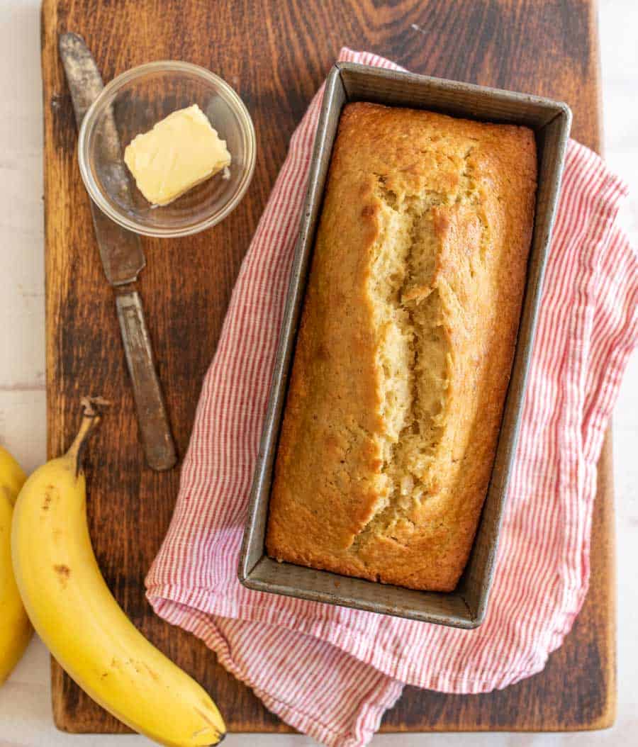 a loaf of banana bread in the pan on top of a striped dish towel, laying on a cutting board with a small dish of butter and a single banana laying to the left side