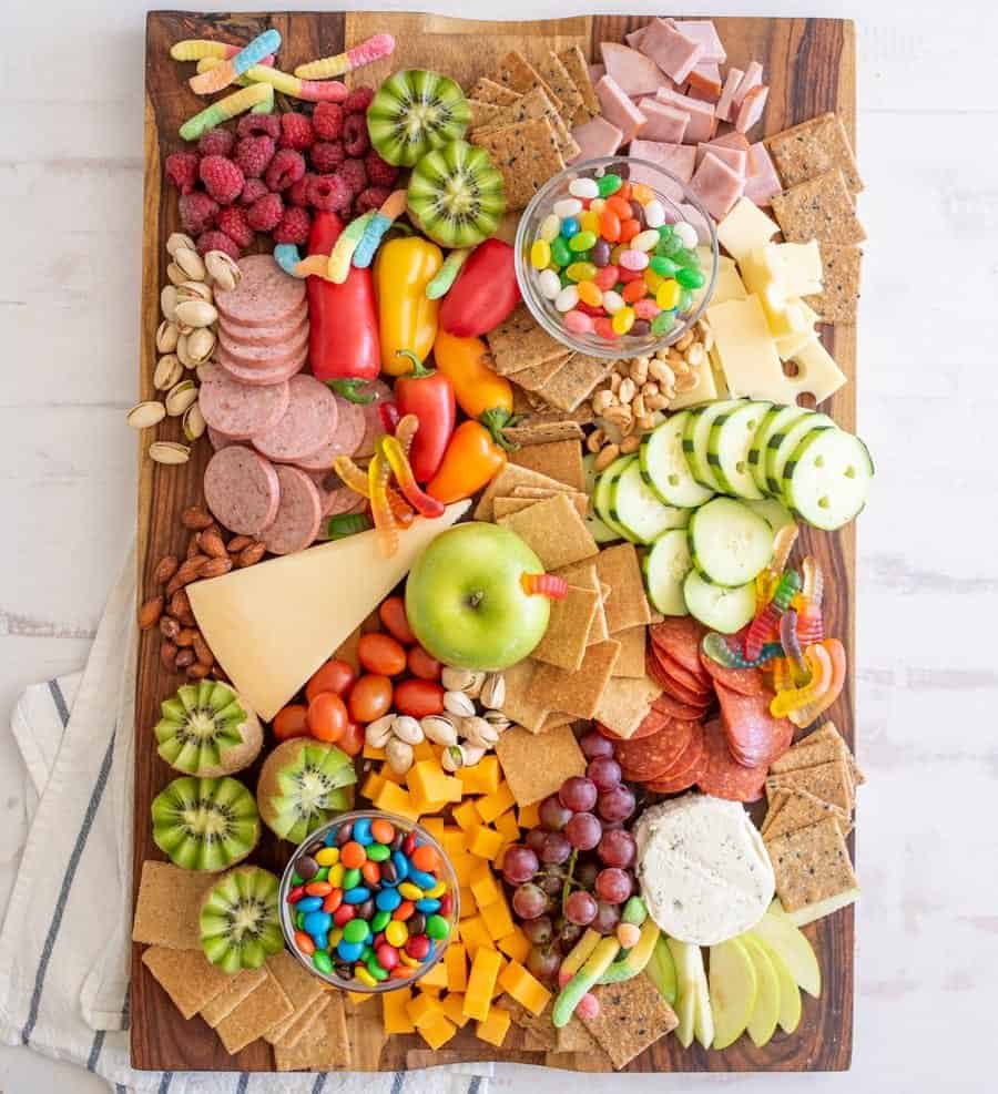 a wooden cutting board full of crackers, cheeses, fruits, veggies, and candies