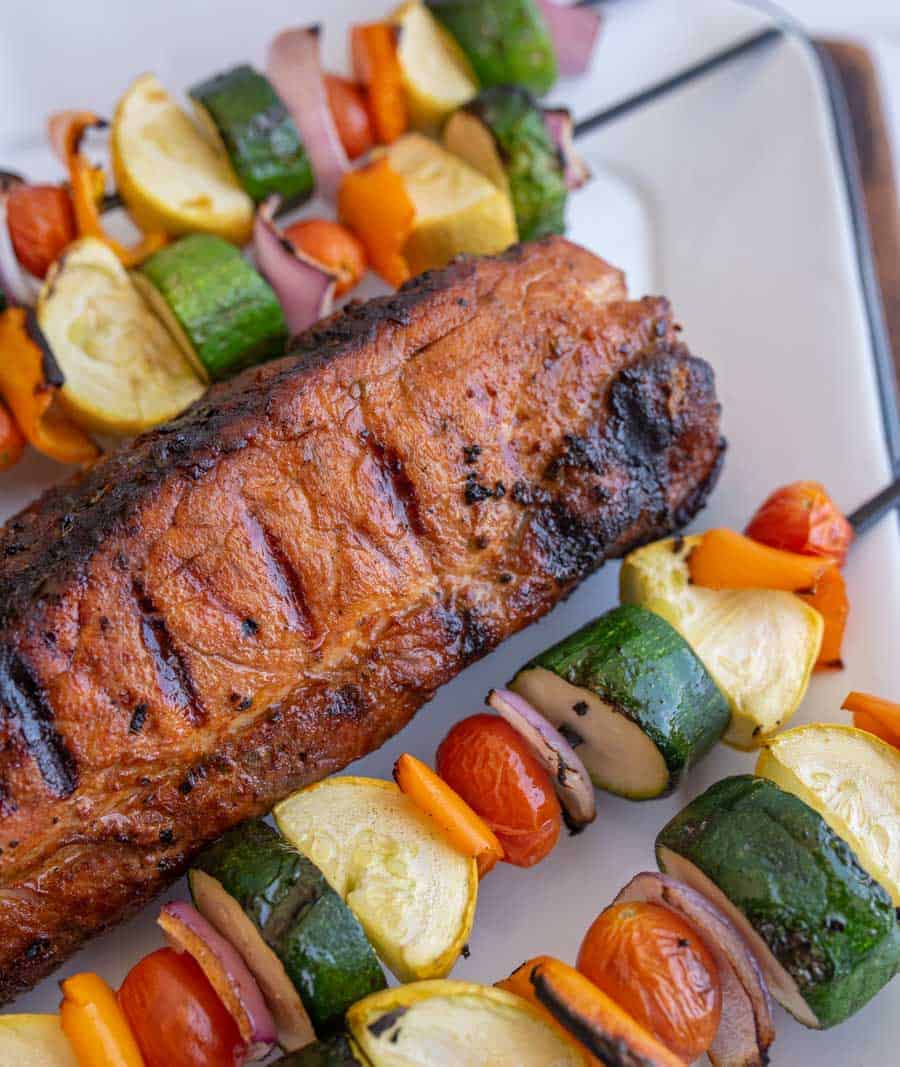 grilled whole pork loin with veggie kabobs on either side on a white serving tray