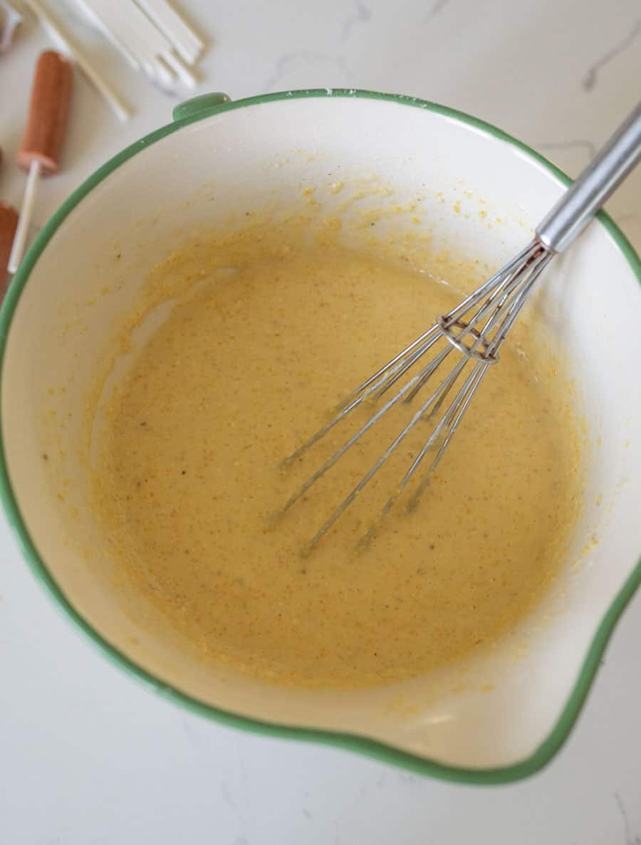 A metal whisk rests in a large white mixing bowl with a green rim, containing thick yellow batter for a corn dog recipe. The bowl sits on a white countertop with popsicle molds visible in the background.