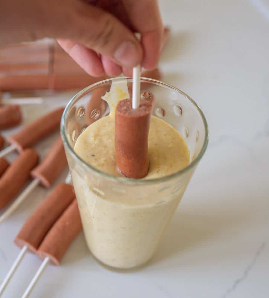 A hand dips a hot dog on a stick into thick batter, following a classic corn dog recipe. Several more hot dogs on sticks rest on the white counter in the background.