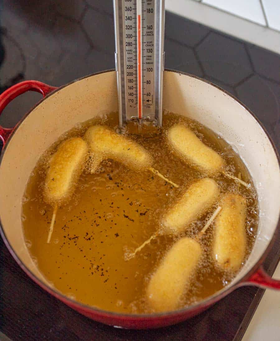 Six corn dogs are being deep-fried in hot oil in a red Dutch oven, with a thermometer clipped to the side for monitoring the oil temperature—perfect for trying out your favorite corn dog recipe.