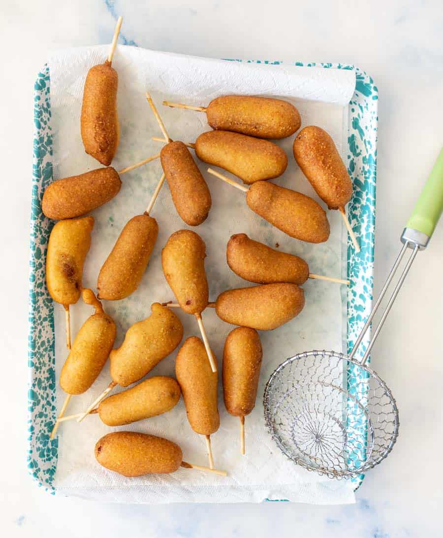 A tray lined with paper towels holds several golden-brown mini corn dogs on sticks, next to a metal strainer with a green handle—perfect results from your favorite corn dog recipe.