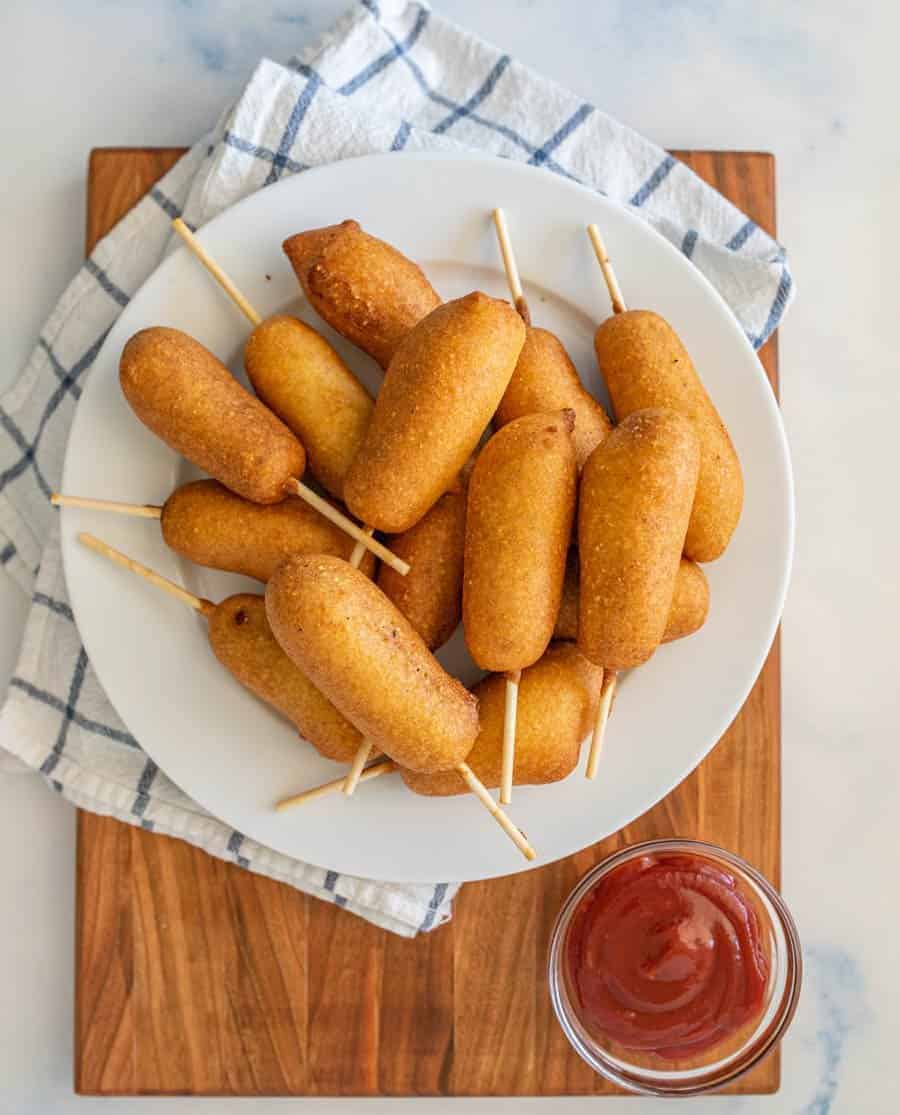 A white plate of mini corn dogs on sticks, made with a classic corn dog recipe, sits on a wooden board with a checkered towel underneath. A small bowl of ketchup is placed next to the plate.