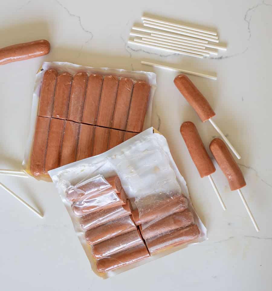 Two open packs of hot dogs on a white surface, with several hot dogs partially inserted onto white sticks—perfect for making homemade corndogs—and extra sticks scattered nearby.