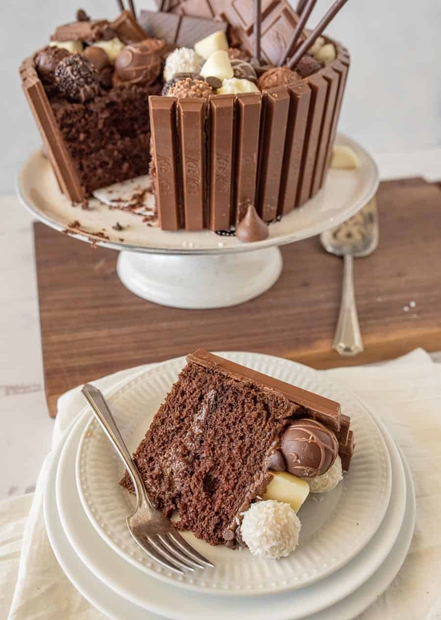 A slice of chocolate candy cake on a plate with a fork and the rest of the cake in the background.