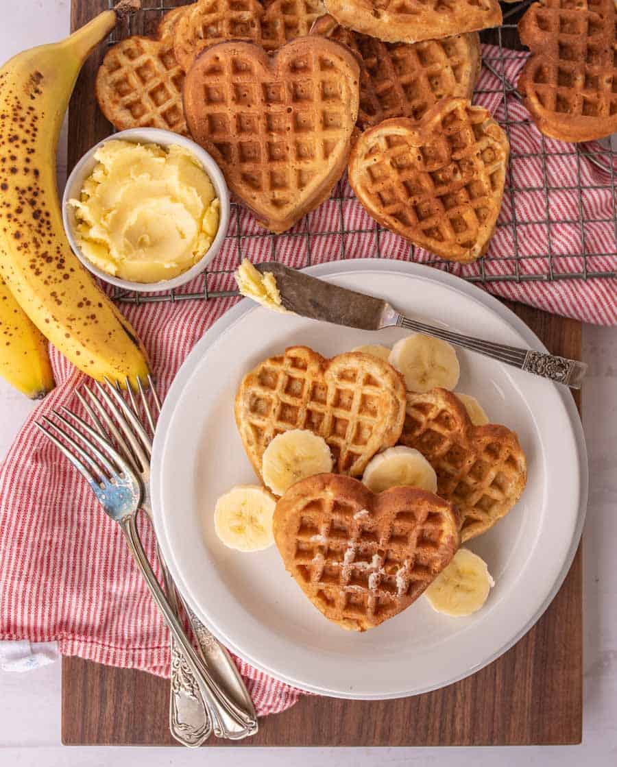 top view of heart shaped waffles and slices of bananas on a plate with utensils, bananas, bowl of butter, and other heart shaped waffles on a cooling rack.