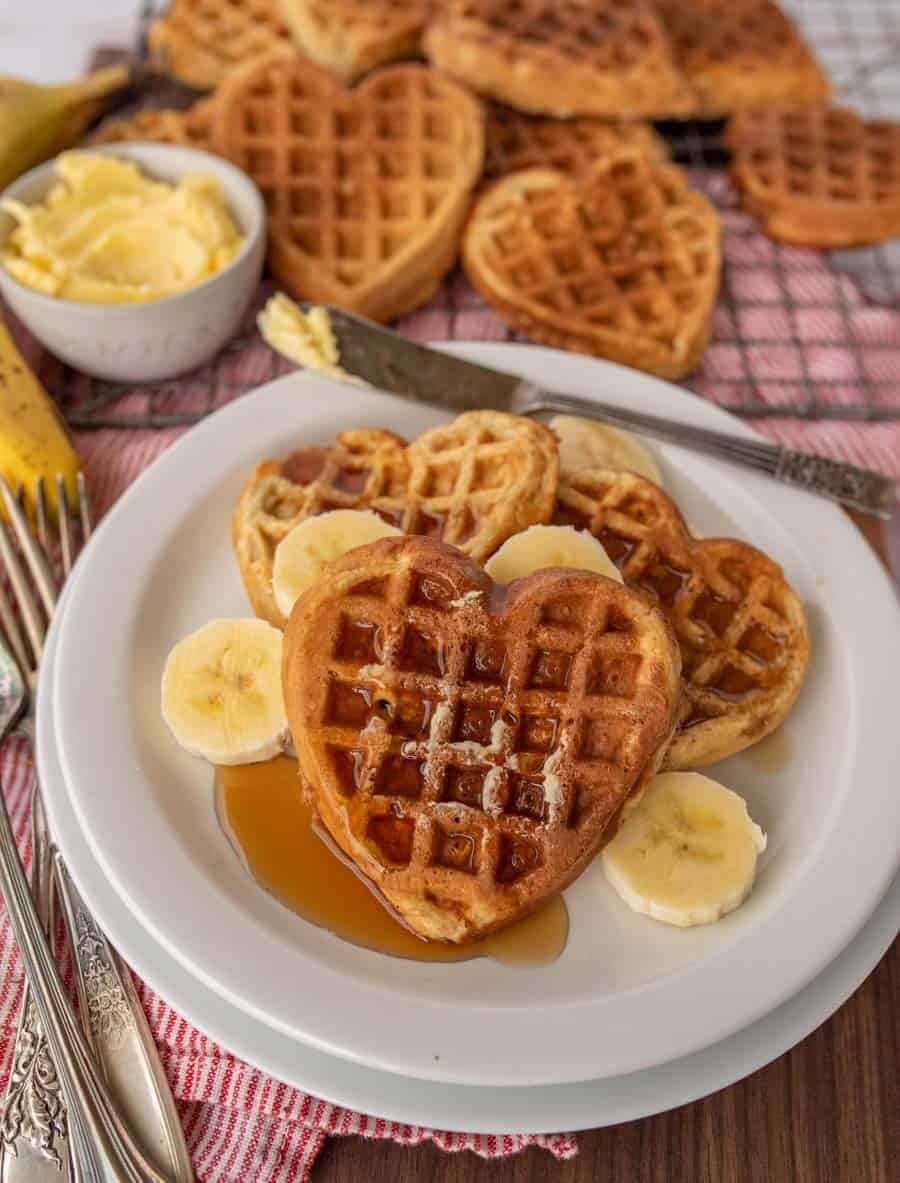 A plate with heart-shaped banana oatmeal waffles topped with sliced bananas and syrup. A bowl of butter sits beside the plate, with more waffles in the background on a checkered cloth. Silverware rests next to the plate on a wooden table.