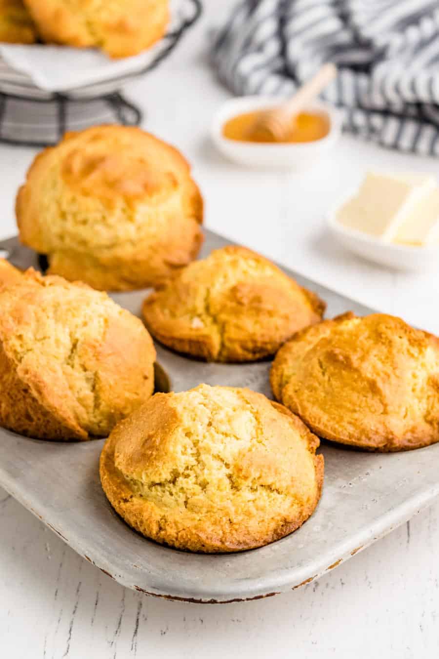 A muffin pan with five golden-brown muffins rests on a white surface, crafted from a classic corn muffin recipe. In the background are a small dish of butter, a bowl of honey with a dipper, and a striped towel.