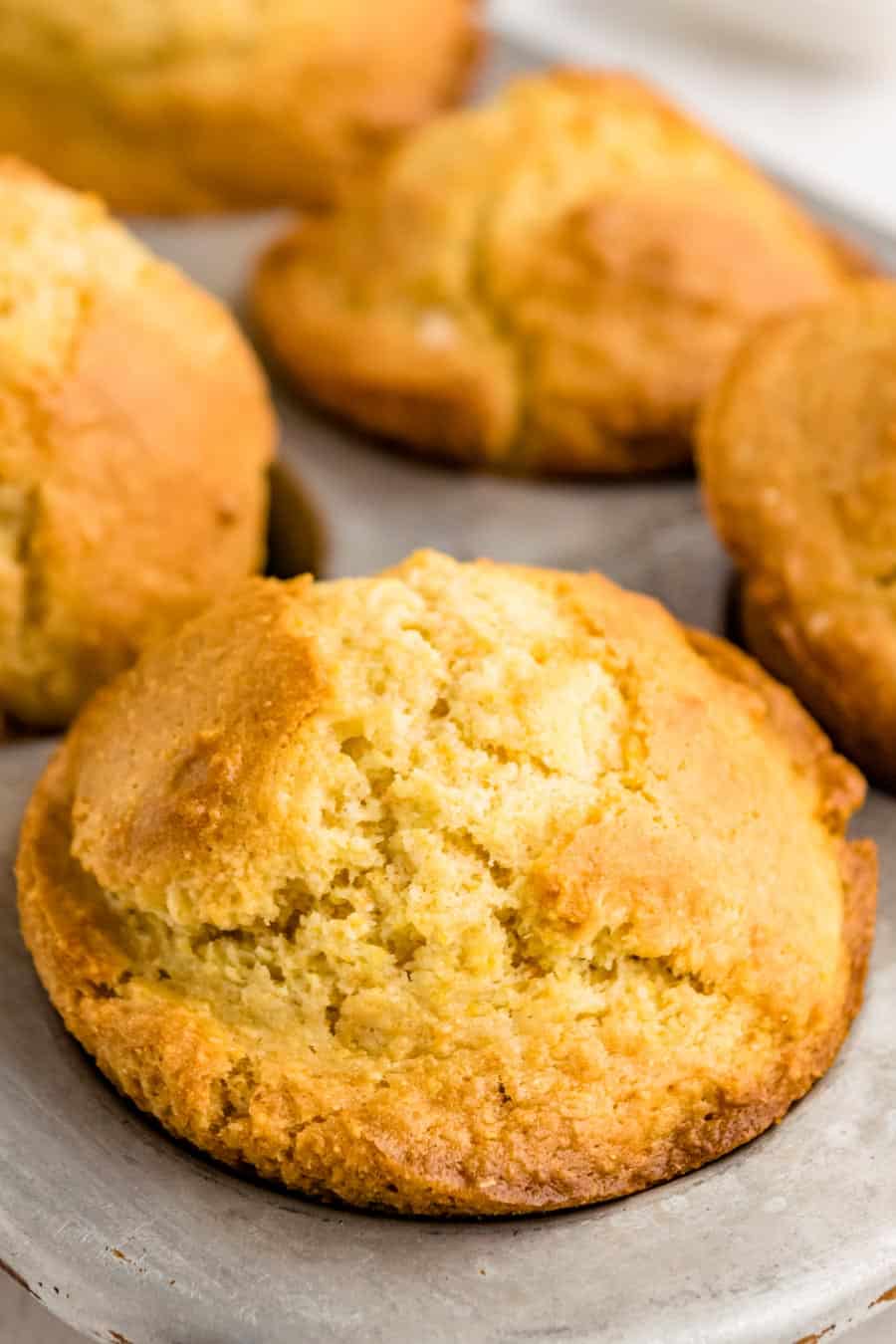 Close-up of golden-brown cornbread muffins with a slightly cracked top, resting in a muffin tin. The texture looks soft and moist—perfect results from your favorite corn muffin recipe, with a warm, freshly-baked appearance.