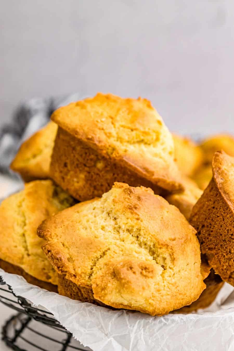 A close-up of several golden brown cornbread muffins, made from a classic corn muffin recipe, stacked on parchment paper and set on a cooling rack with a light, neutral background.