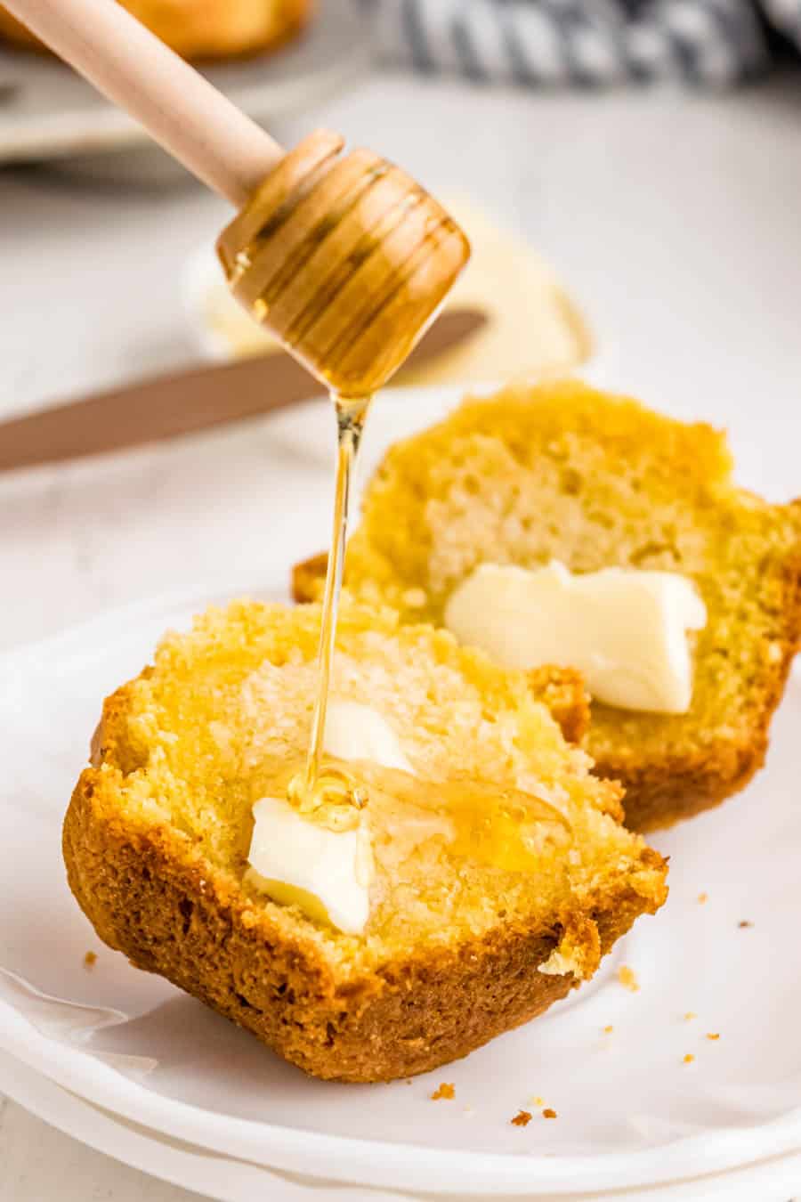 A honey dipper drizzles honey onto two halves of cornbread, each topped with melting butter, on a white plate—perfect inspiration for your next corn muffin recipe.