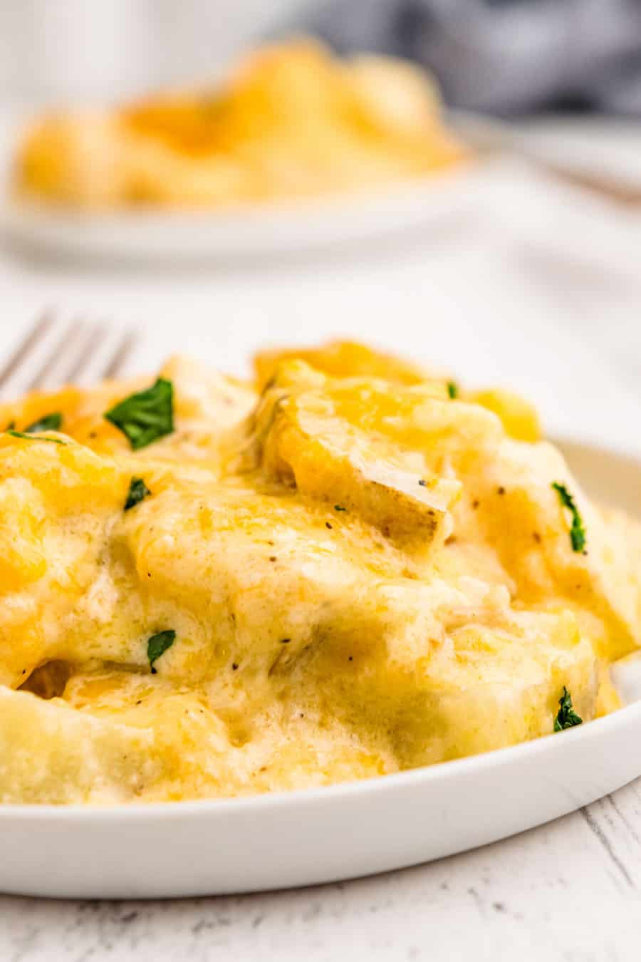A close-up of creamy, cheesy Pioneer Woman's Potatoes au Gratin garnished with parsley, served on a white plate with another plate of food blurred in the background.