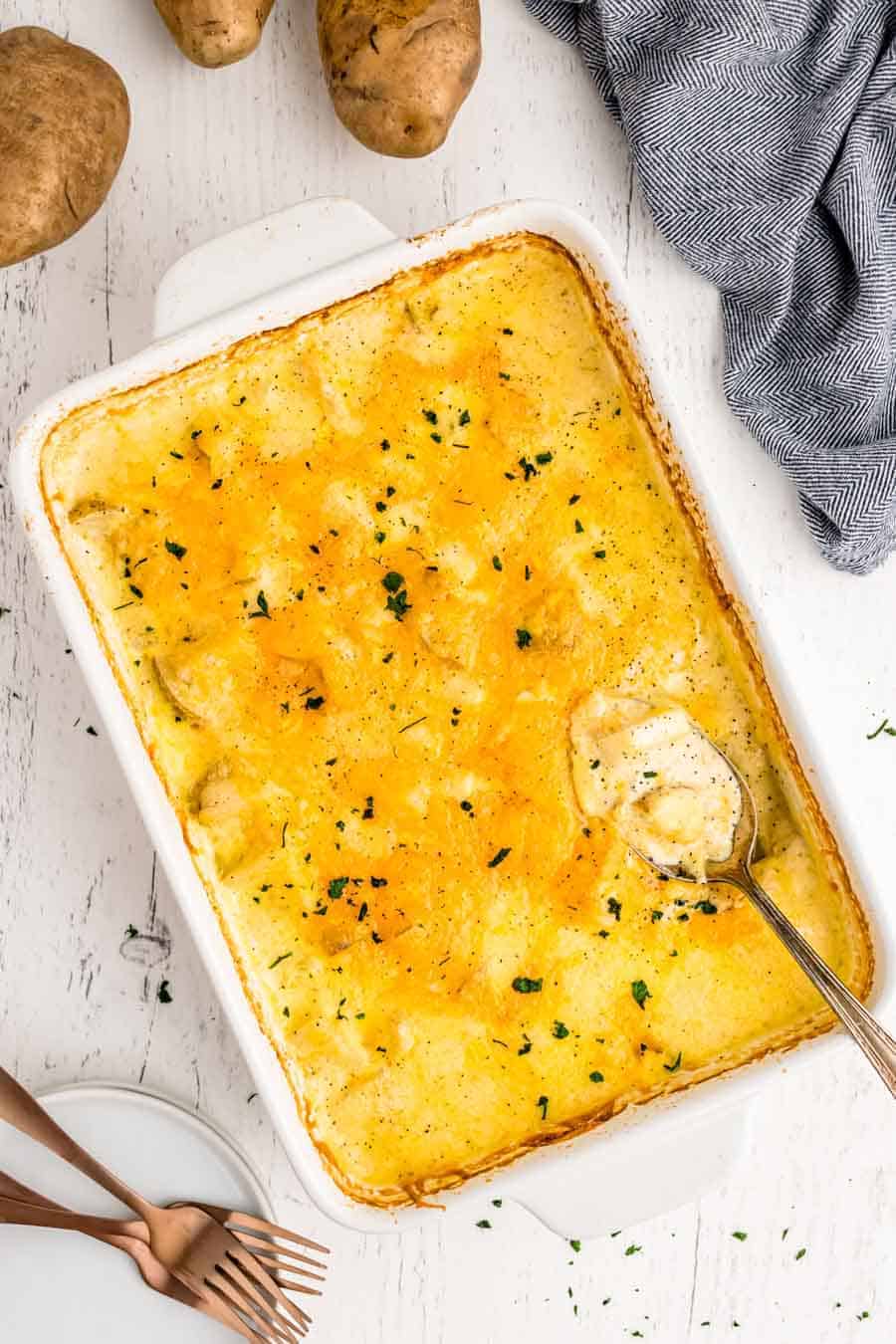 A baked casserole dish filled with cheesy scalloped potatoes, garnished with herbs. A serving spoon rests in the creamy, golden layers, with some potatoes scooped out. Nearby are whole potatoes, a gray cloth, plates, and forks.