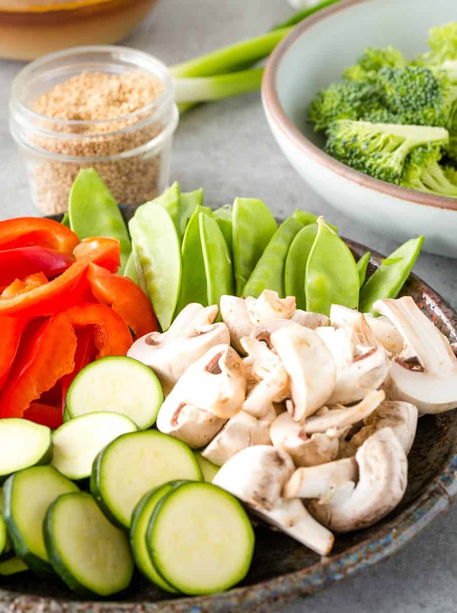 A bowl of cut up mushrooms, zucchini, bell pepper, and snow peas with a bowl of broccoli in the background.