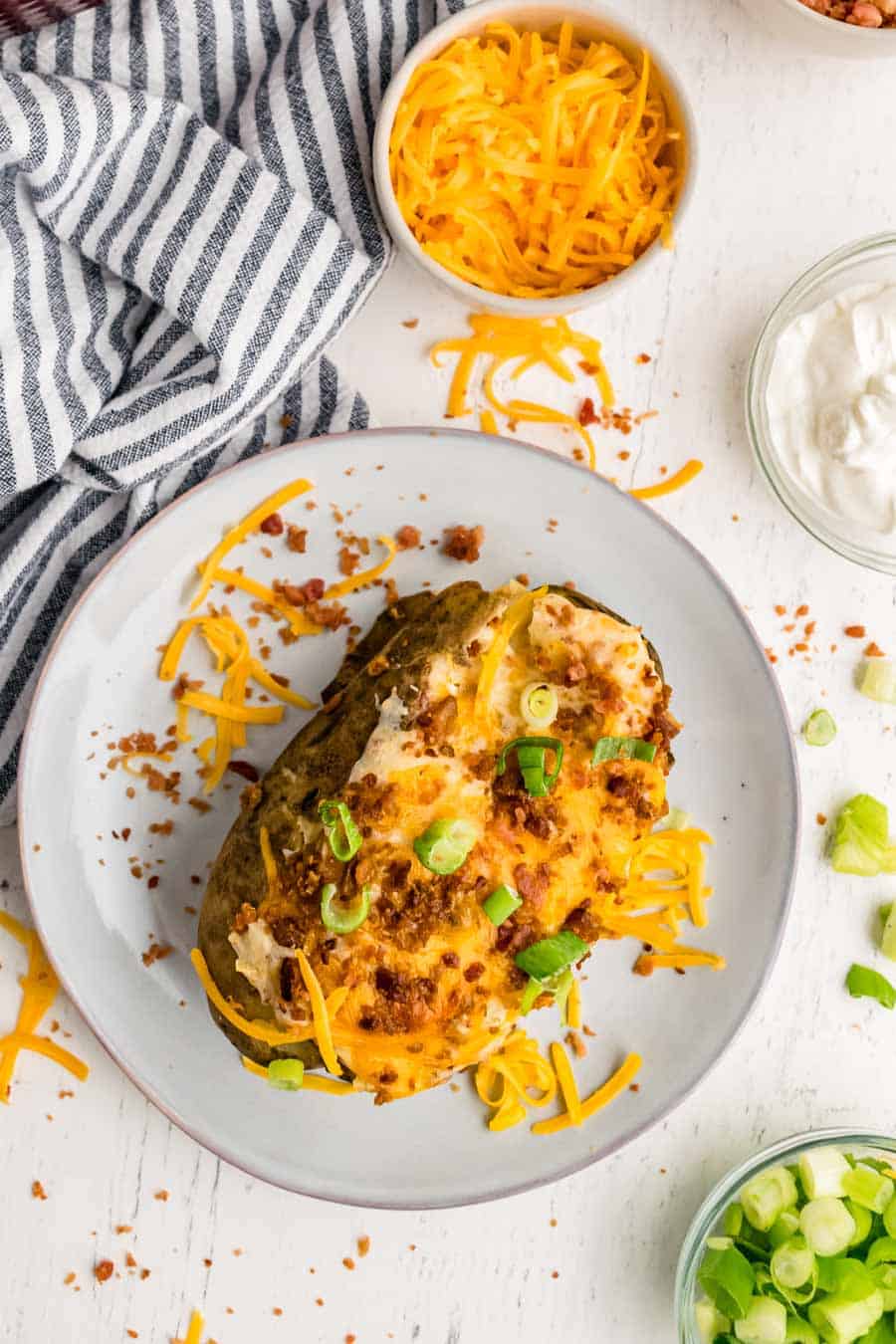 Top view of a Twice Baked Potatoes topped with cheese, green onions, and bacon on a white plate with bowls of cheese, sour cream, and green onions on the side.