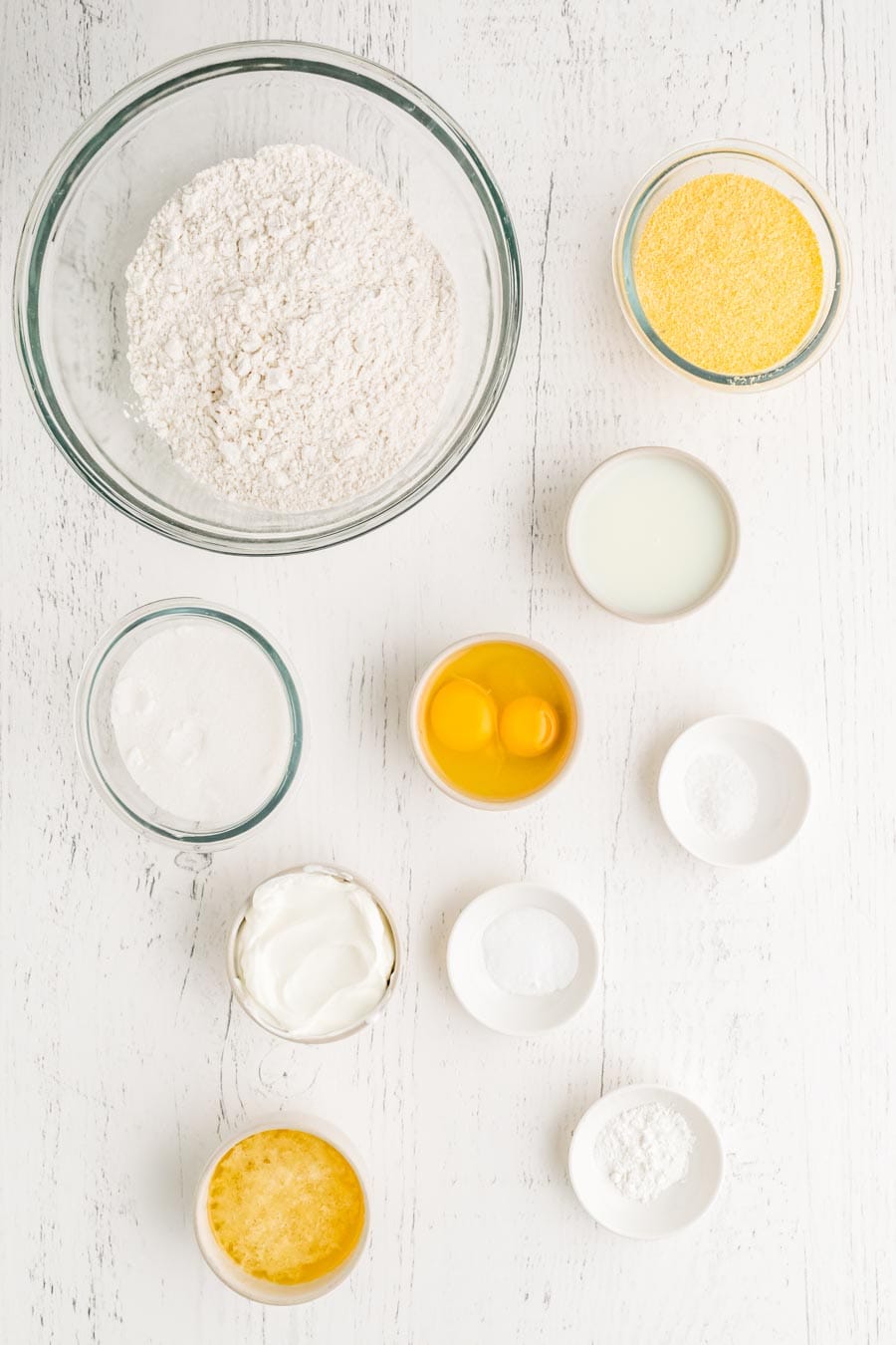 A variety of baking ingredients in separate bowls on a white wooden surface, including flour, cornmeal, eggs, sugar, and more—everything you need for a homemade corn muffin recipe.