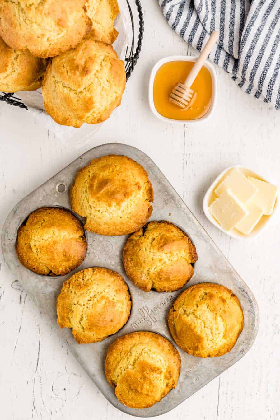 A muffin tin with six golden brown muffins from a delicious corn muffin recipe sits on a white surface beside a wire basket of more muffins, a dish of honey with dipper, butter slices on a plate, and a striped cloth napkin.
