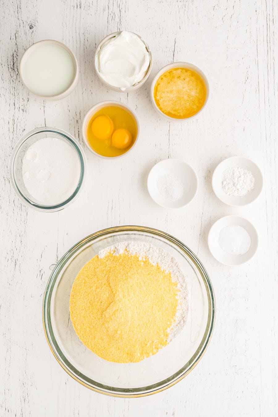 A top-down view of baking ingredients for a corn muffin recipe on a white surface, featuring bowls with cornmeal, sugar, eggs, milk, sour cream, melted butter, and small dishes with salt, baking powder, and baking soda.