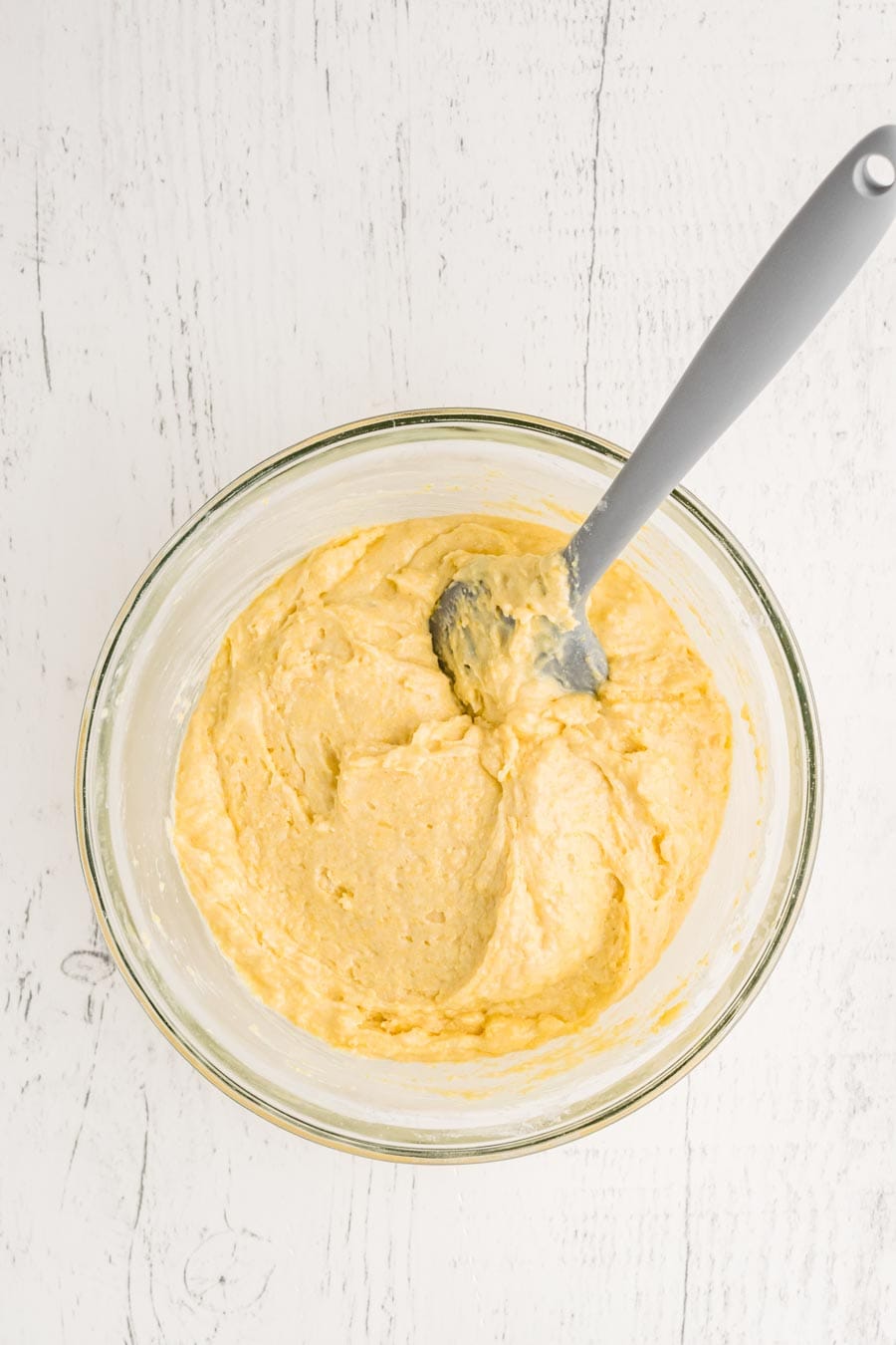 A glass bowl filled with thick, pale yellow batter for a corn muffin recipe and a gray-handled spoon, set on a white wooden surface.