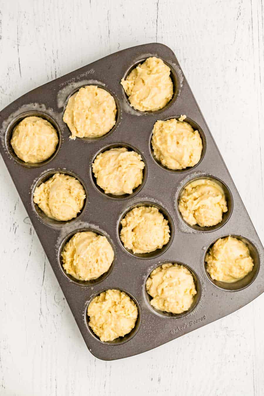 A muffin tin filled with 12 portions of unbaked corn muffin recipe batter, placed on a white wooden surface.