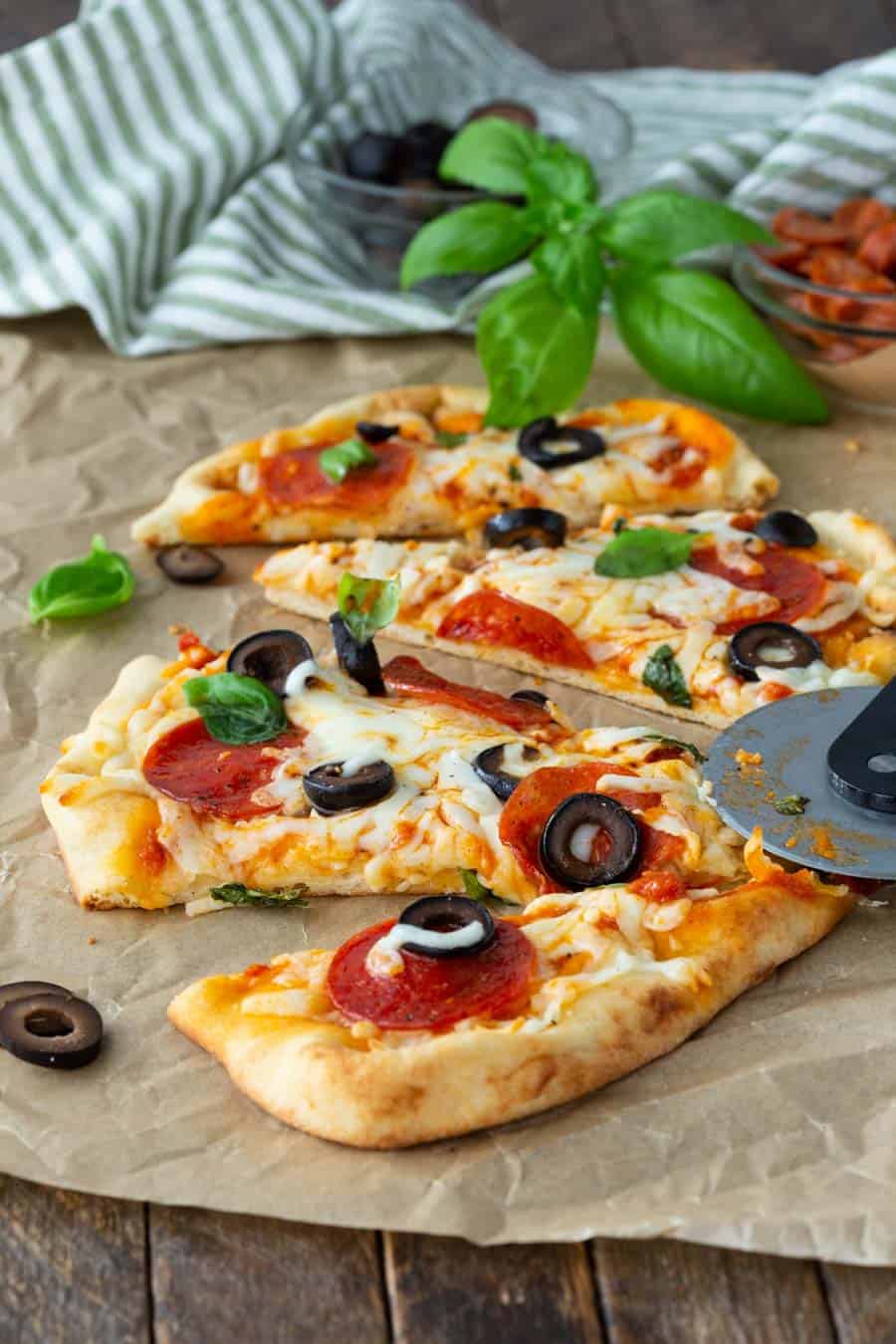 Recipe for a flatbread pizza recipe with a sliced pizza on parchment paper, topped with pepperoni, black olives, and melted cheese, showcases an easy pepperoni flatbread pizza recipe. A pizza cutter is nearby, with a striped cloth and fresh basil leaves adding a touch of charm in the background.