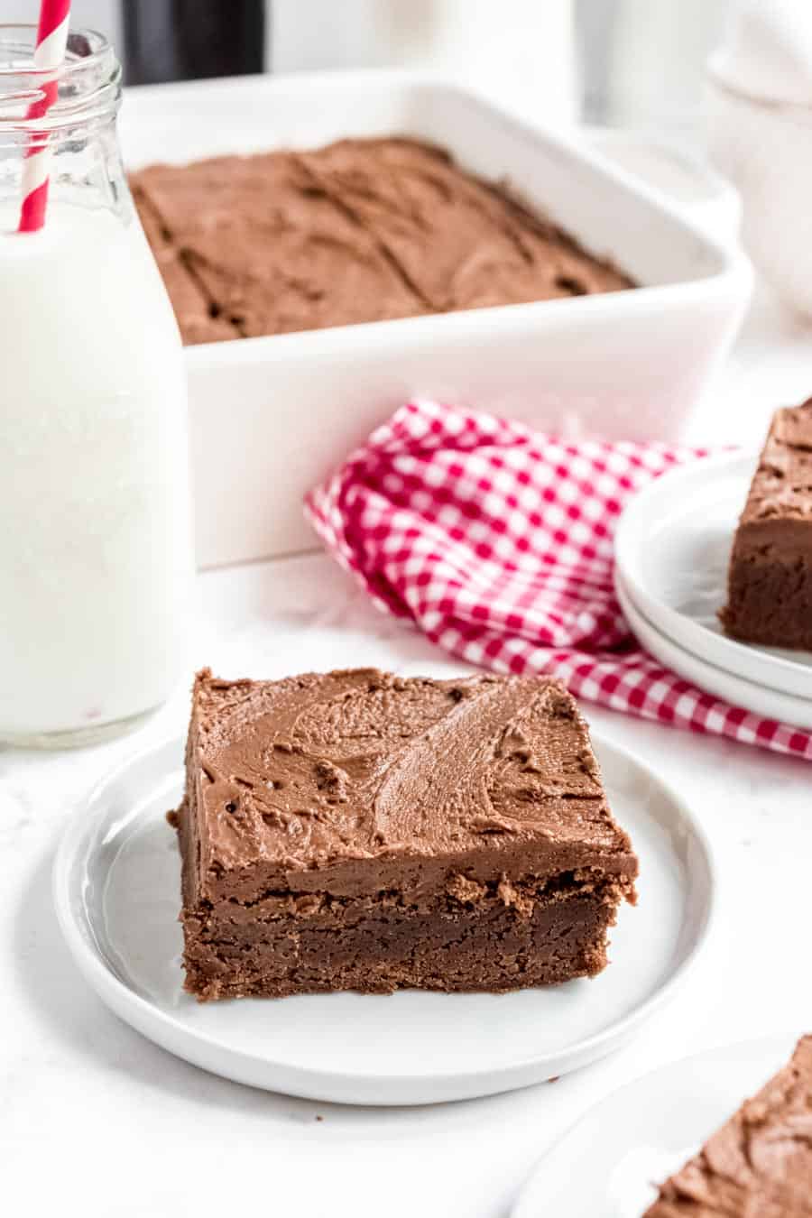 A brownie square with frosting on a white plate with milk in the background.