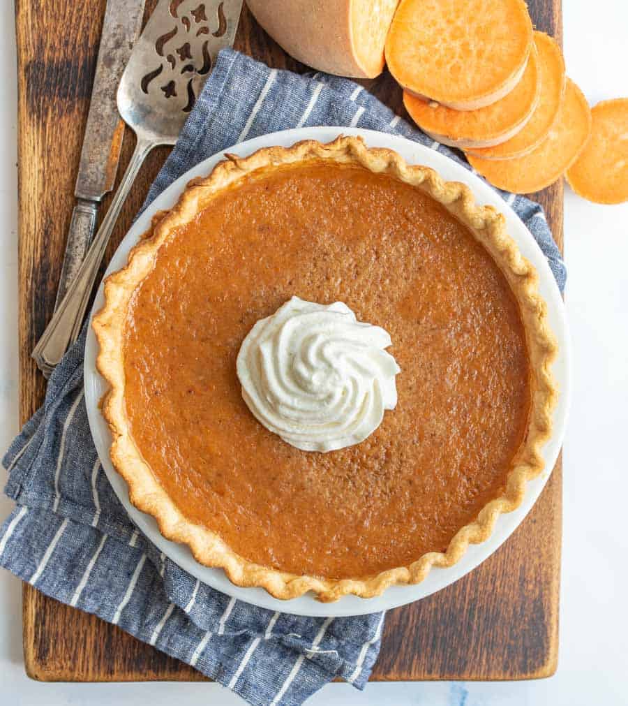 Top view of a sweet potato pie topped with whipped cream with serving utensils and slices of sweet potatoes.