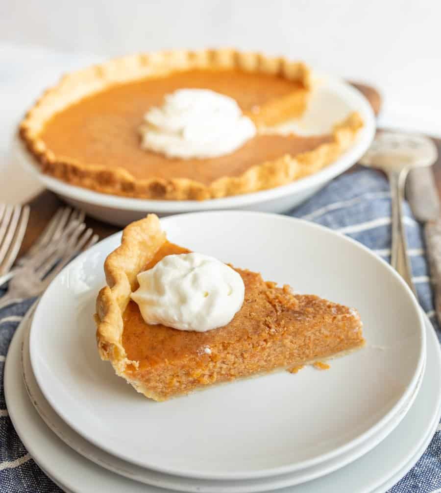 A slice of sweet potato pie with whipped cream on top with the rest of the pie in the background.