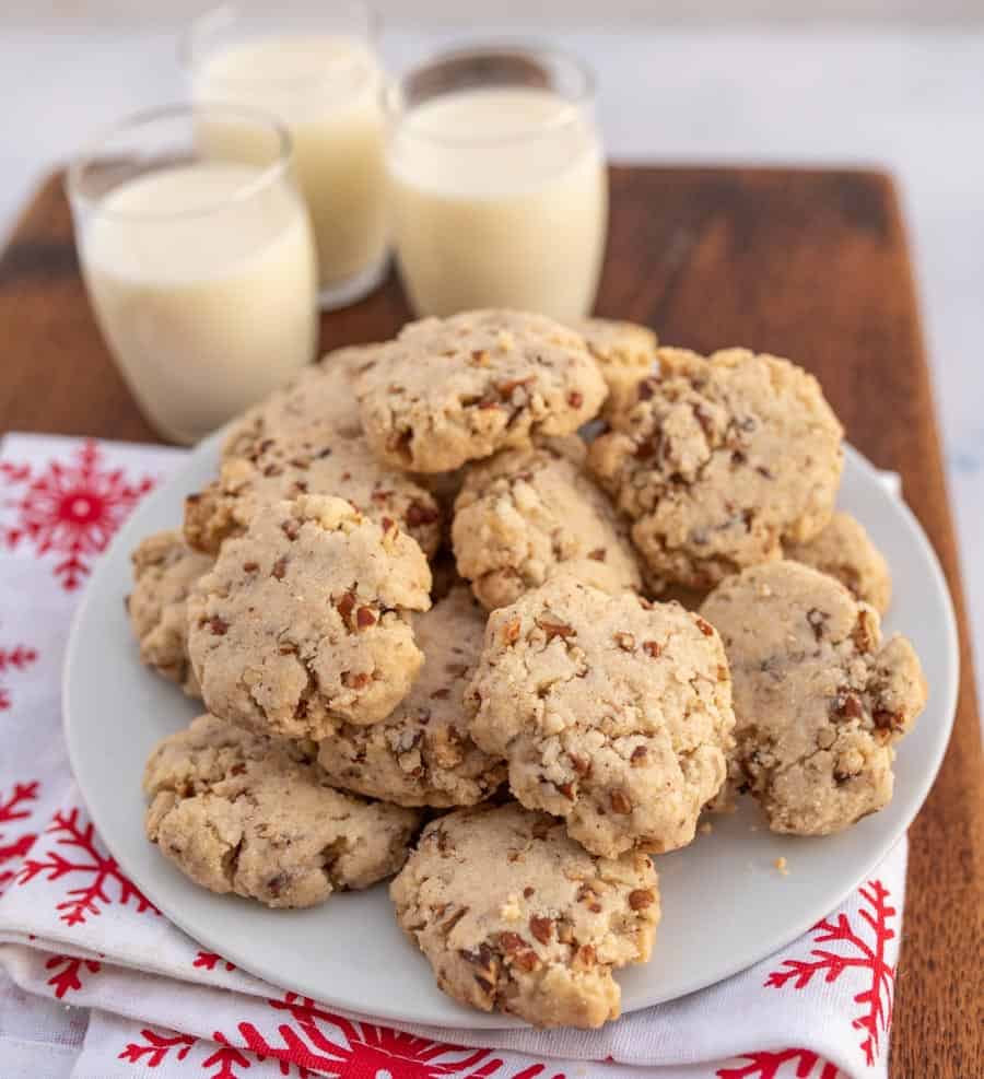 A Christmas setting on wooden block with egg nog and traditional tan looking pecan sandies cookies on a white plate.
