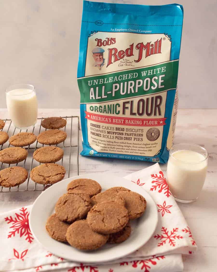 Ginger snap cookies on a white plate on a white linen with red snowflakes. and baking rack with glasses of milk and a bag of flour.