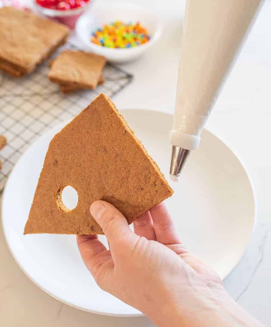 A hand holding a gingerbread house wall about to ice with royal icing in a pipet.