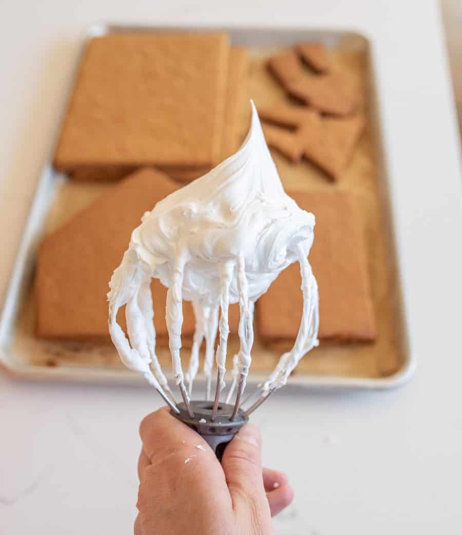 a mixing whisk with peaked white icing held out in front with deconstructed gingerbread house walls and cookie pieces on a sheet pan in the background
