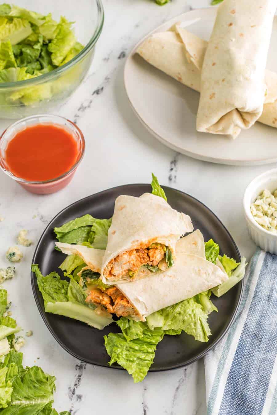 Who halves of a buffalo chicken wrap stacked on a bed of lettuce on a black plate with hot sauce and more wraps in the background.