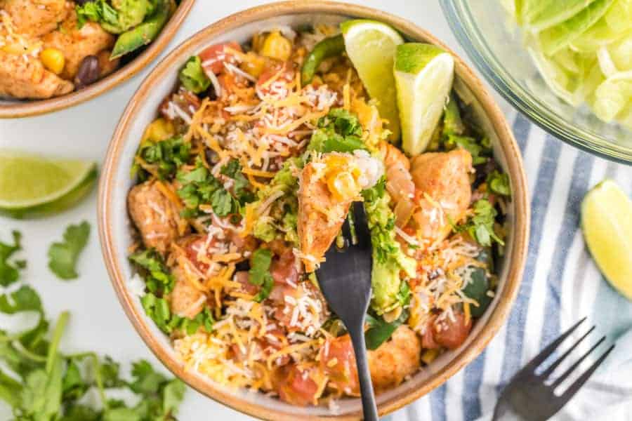 A very close view of chicken burrito bowl to see detail of limes and cilantro and cheese avocados and tomatoes with fork.