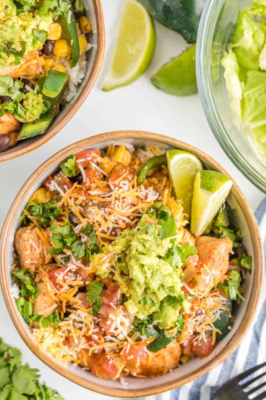 Top view of a chicken burrito bowl with lettuce, tomatoes, cheese, guacamole, cilantro, and limes.
