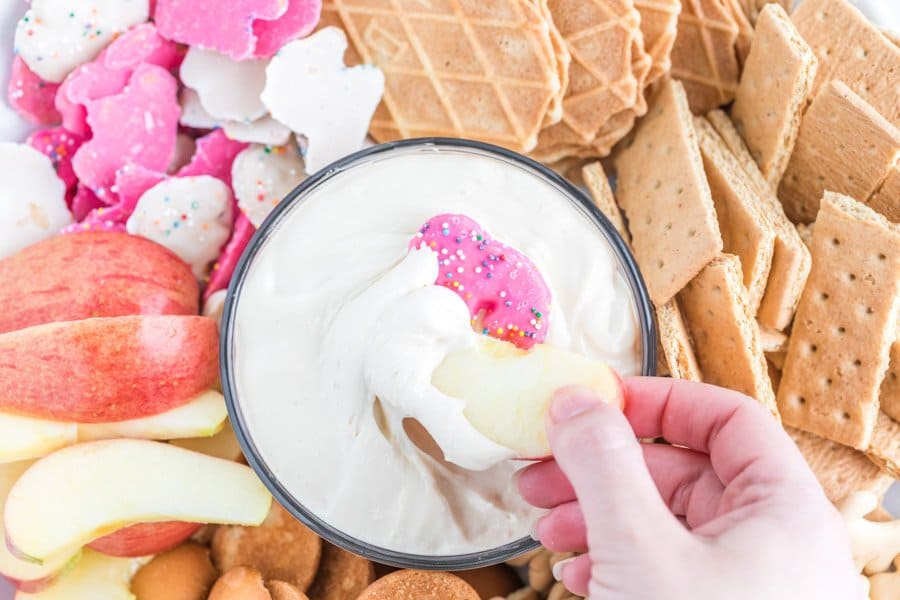 POV of apple in cheesecake dip - top view of plated snacks and other dip-able foods such as gram crackers, apples, and wafers arranged around a glass bowl with a silky white cheesecake dip with a pink iced circus animal in the center.