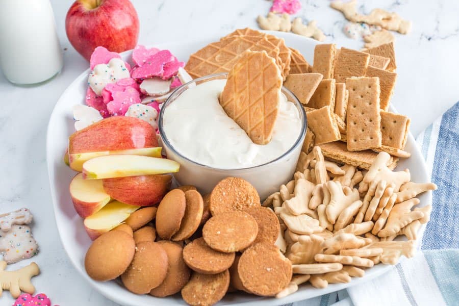 Side view of a white platter with cookies and fruit with a bowl of cheesecake dip in the center with a wafer dipped in it.