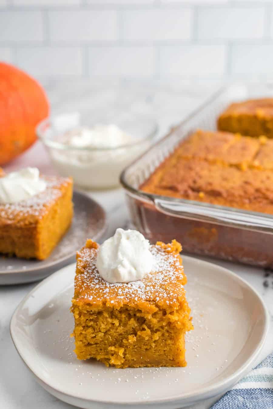 A square servings of extra thick pumpkin cake with whipped cream on top on white plates next to the baking dish.