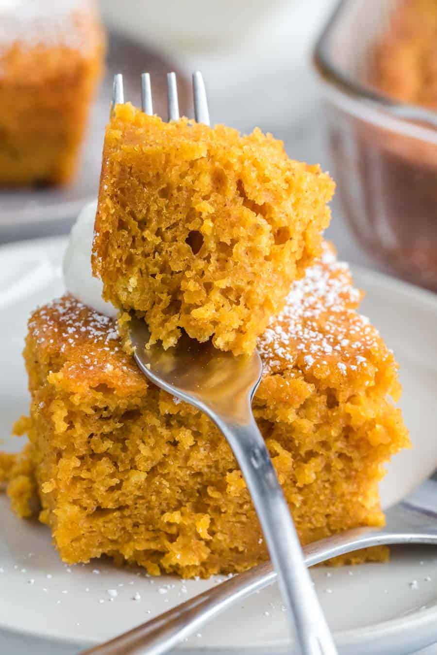 Close up POV of a square serving of extra thick pumpkin cake on a white plate with a dollop of cream and one bite on a fork closest to the camera lens.
