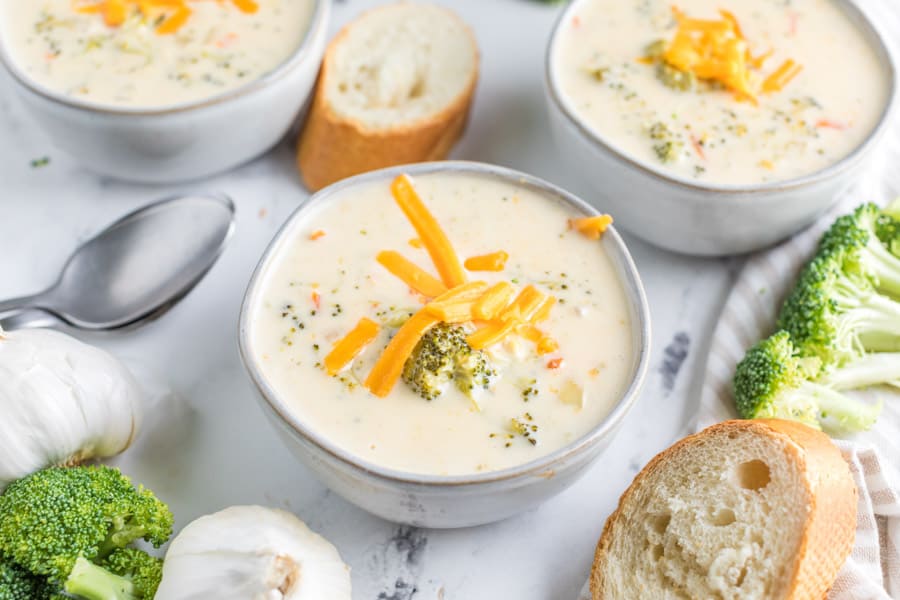 Three bowls of broccoli and cheese soup with cheese sprinkles on top and slices of bread next to the bowls.