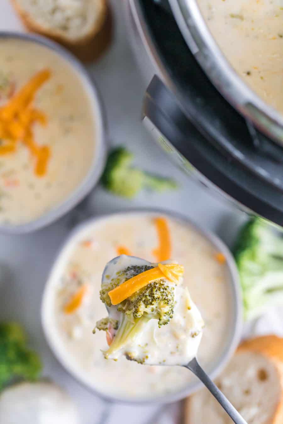 POV of a spoon full of soup over a small white ceramic cup of creamy broccoli cheddar soup with specks of seasoning and cooked broccoli garnished with shredded cheddar cheese.