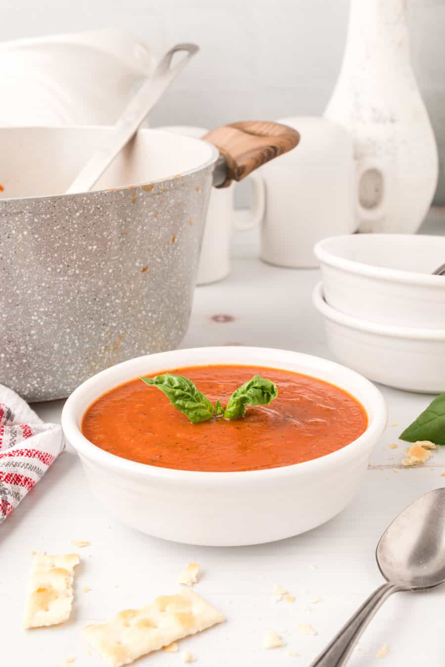 A bowl of roasted tomato soup with basil on top next to the pot of soup.