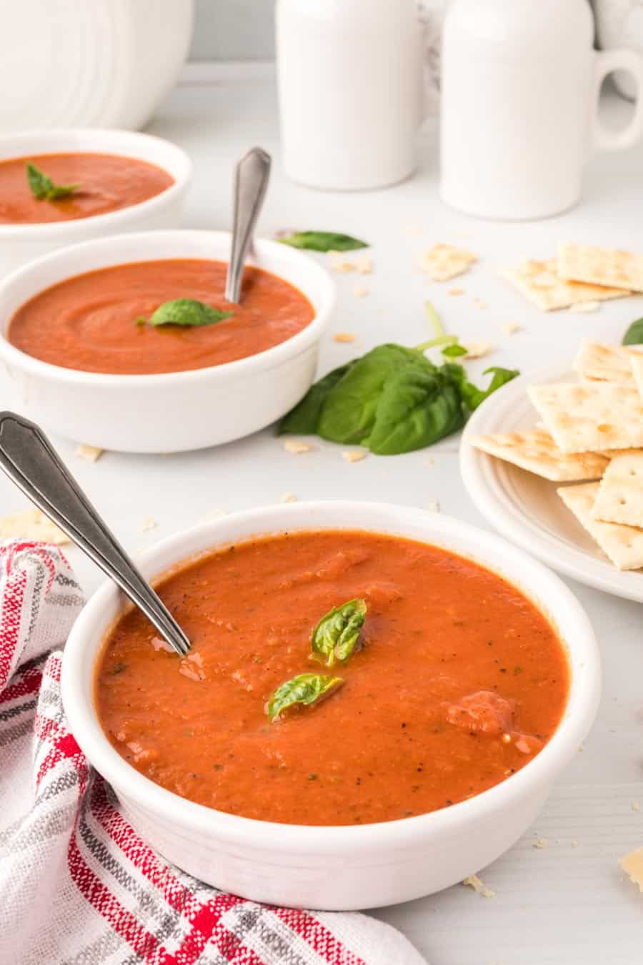 A bowl of tomato basil soup with basil leaves on top and a spoon in it next to two other bowls of soup.