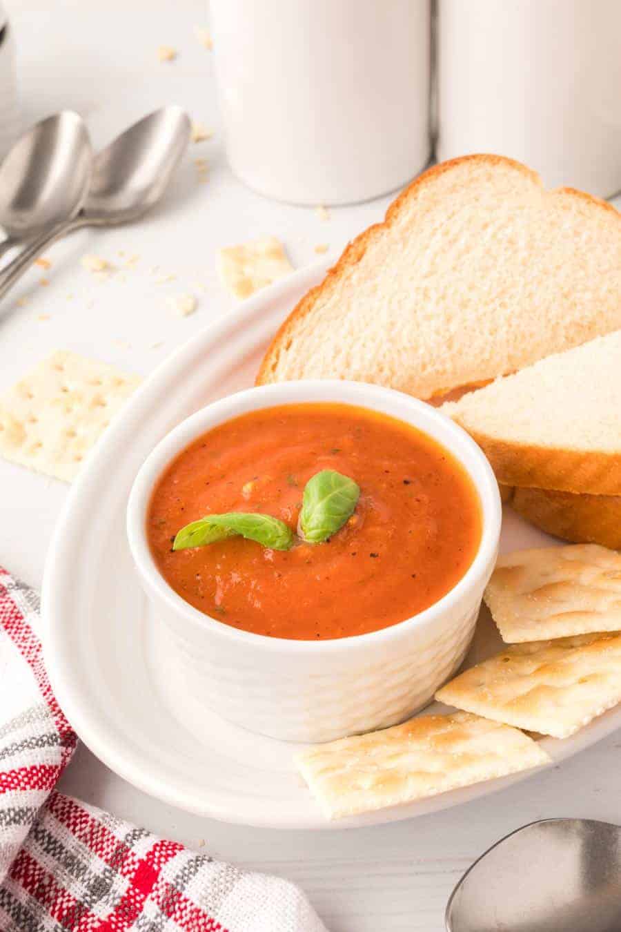A white bowl of roasted tomato soup with basil leaves on top on a plate with Saltines and white bread.