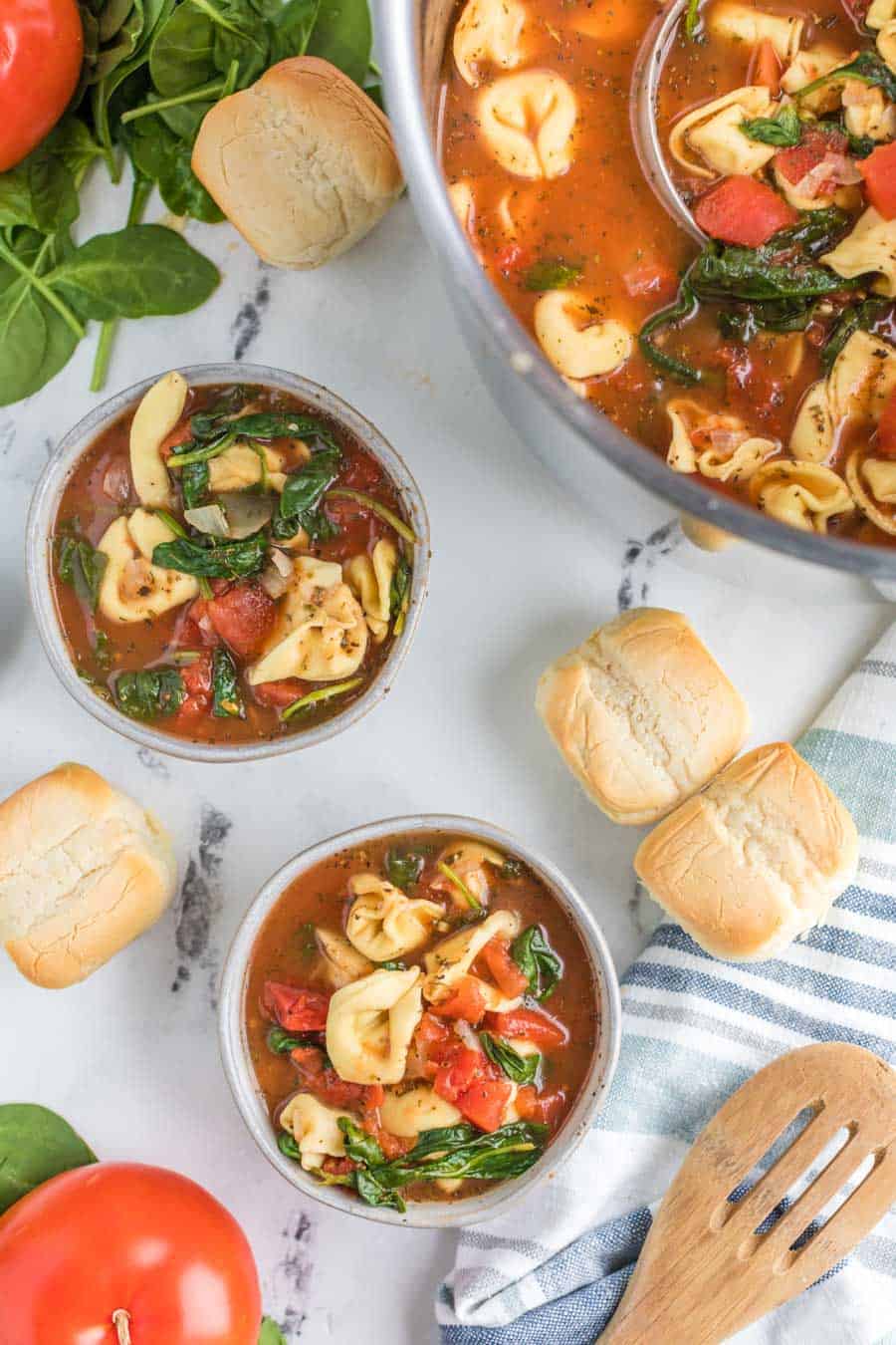 Top view of bowls of tortellini tomato soup in a scene over marble table with rolls, whole tomatoes, a wooden spoon, and the edge of the large pot of soup with a ladle in it.