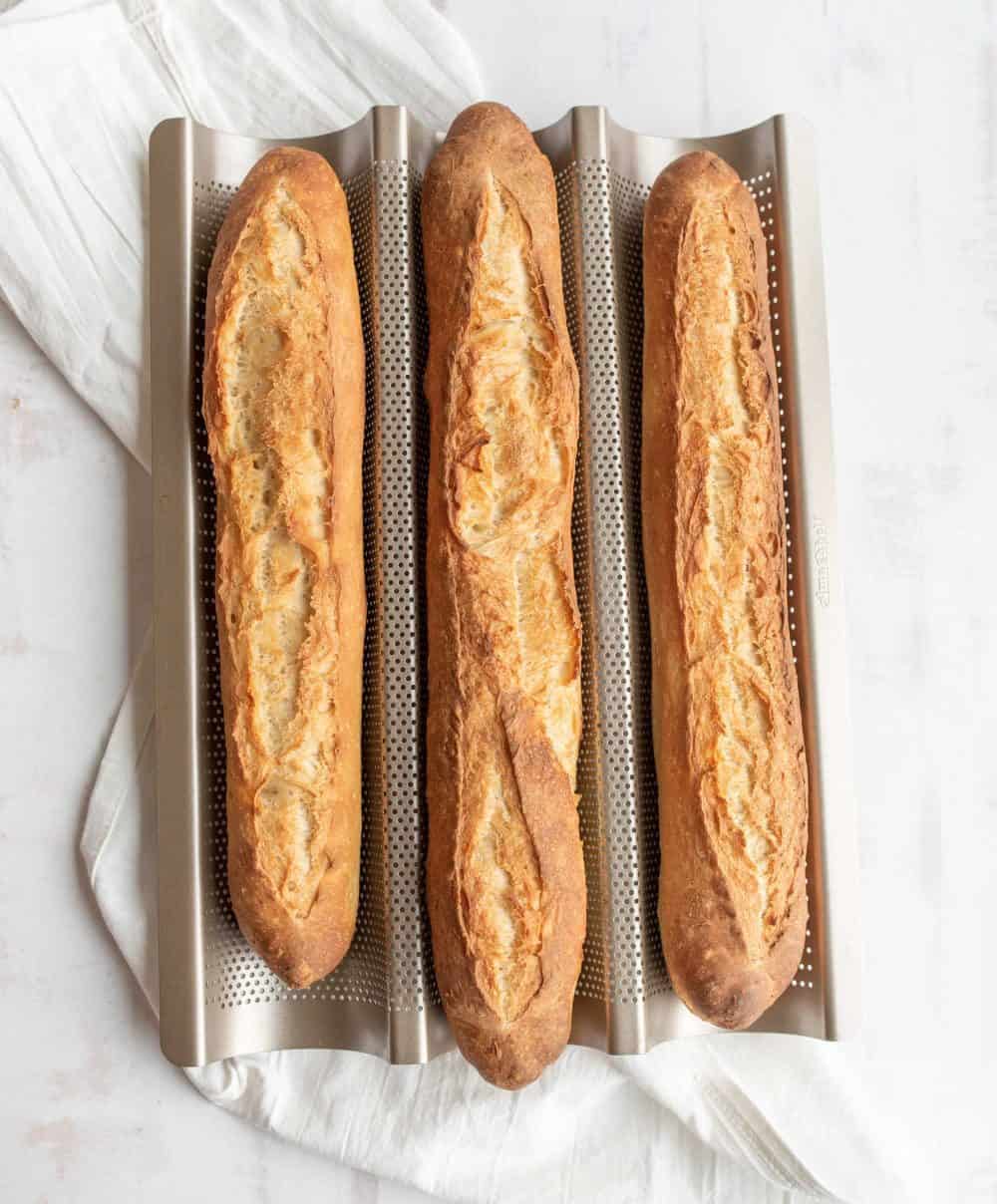 Three freshly baked baguettes, made from a classic baguette recipe, rest on a perforated baking tray atop a white cloth and light surface.
