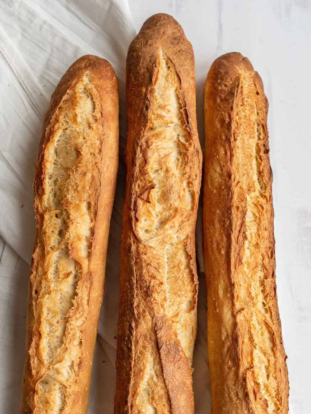 Three golden-brown baguettes, made from a classic baguette recipe, with crisp, textured crusts rest side by side on a white surface, partially overlapping a white cloth.